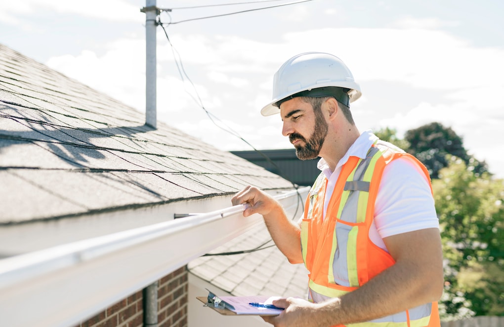 A man in a hard hat, holding a clipboard, performing a roof inspection
